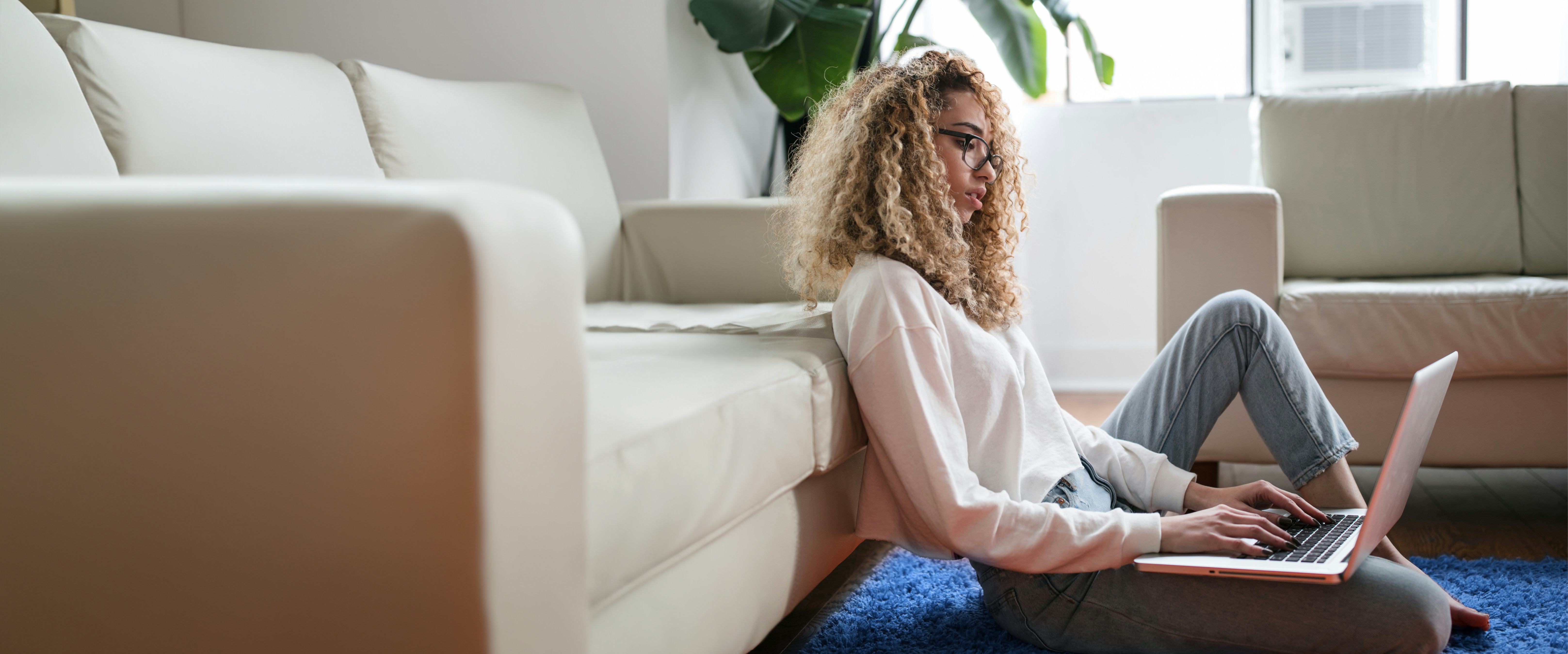 Junge Frau mit blonden locken sitzt auf dem Teppich in ihrer Wohnung und arbeitet an einem Laptop